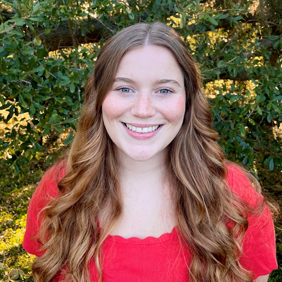 Kaylee Hartsing, Jr. Environmental Scientist, stands wearing business casual attire in front of a leafy green background.